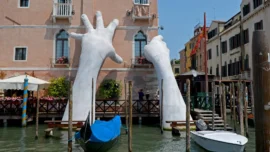 installazione d'arte a venezia che raffigura due braccia che sorreggono un palazzo sulla laguna